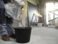 Budowlaniec wsypuje nierówny chudziak pod styropian do czarnego wiadra. W tle widać plac budowy.