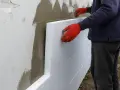 Pracownik w czerwonych rękawiczkach przykłada styropian do ściany. Zatapianie siatki na styropianie cena za metr.