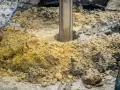 Wiertnica zagłębia się w ziemię, tworząc otwór pod fundamenty palowe. Wokół rozsypana glina i kamienie.