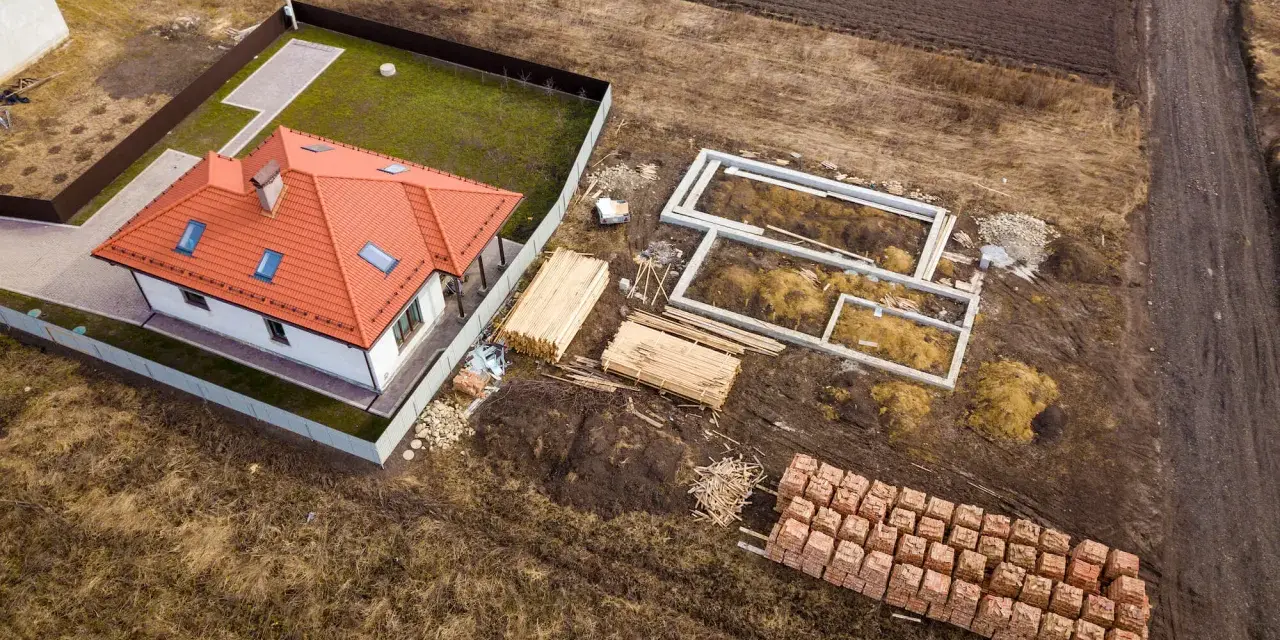 Widok z lotu ptaka na budowę domu. Widać fundamenty, sterty drewna i cegieł, a także wydzielenie drogi z działki rolnej.