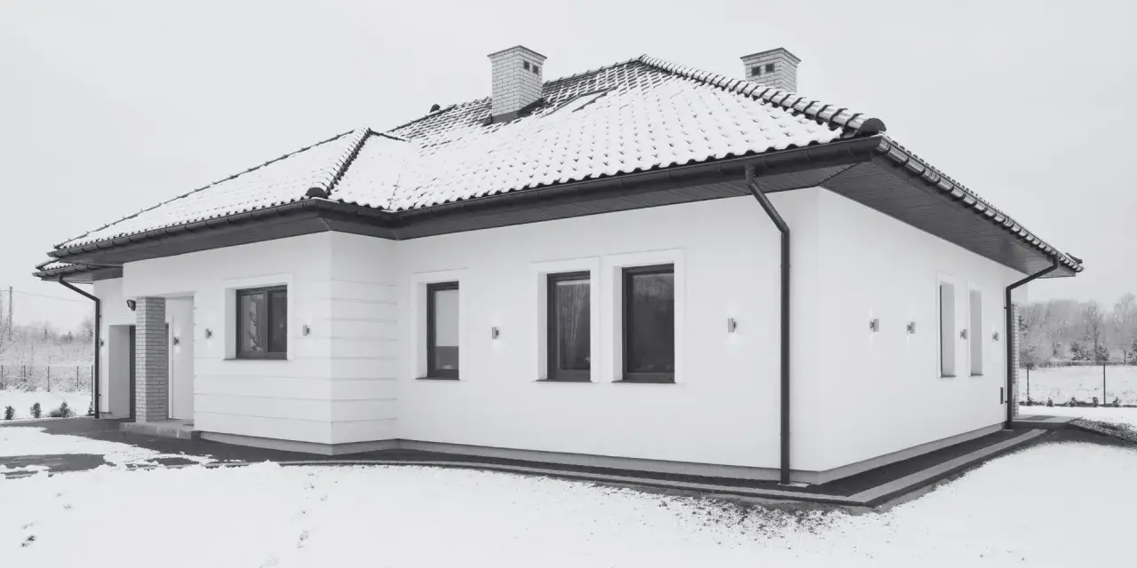 Nowoczesny, biały budynek wolnostojący z dachem pokrytym śniegiem. Wokół domu leży śnieg, tworząc zimową scenerię.
