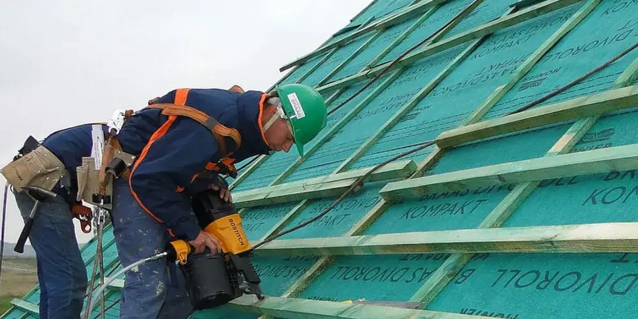 Dwaj dekarze pracują nad dachem, montując tymczasowe zabezpieczenie dachu. Jeden z nich używa pneumatycznego gwoździarki.