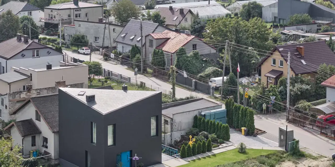 Nowoczesny, czarny dom o trójkątnym kształcie, otoczony innymi budynkami i zielenią. Widać ulicę z samochodami i ludzi. To przykład nowoczesnego użytek ba.