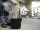 Budowlaniec wsypuje nierówny chudziak pod styropian do czarnego wiadra. W tle widać plac budowy.