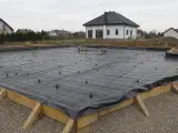 Folia pod ławy fundamentowe rozłożona na placu budowy, zbrojenie gotowe do zalania betonem. W tle dom w budowie.