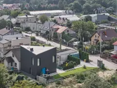 Nowoczesny, czarny dom o trójkątnym kształcie, otoczony innymi budynkami i zielenią. Widać ulicę z samochodami i ludzi. To przykład nowoczesnego użytek ba.