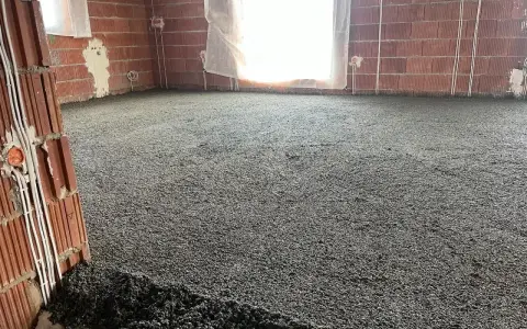 Wylewki styrobeton na podłodze w budowie. Ściany z cegły, okna zasłonięte, widoczne instalacje elektryczne.