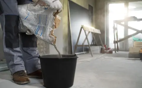 Budowlaniec wsypuje nierówny chudziak pod styropian do czarnego wiadra. W tle widać plac budowy.