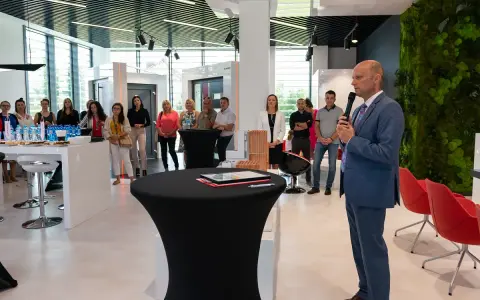Mężczyzna w garniturze przemawia do grupy osób w nowoczesnym salonie okien i drzwi.