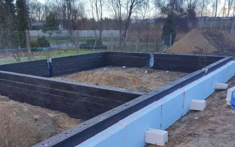 Budowa domu w toku: wylewane fundamenty z izolacją, widoczne zbrojenie i piasek.