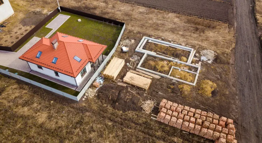 Widok z lotu ptaka na budowę domu. Widać fundamenty, sterty drewna i cegieł, a także wydzielenie drogi z działki rolnej.