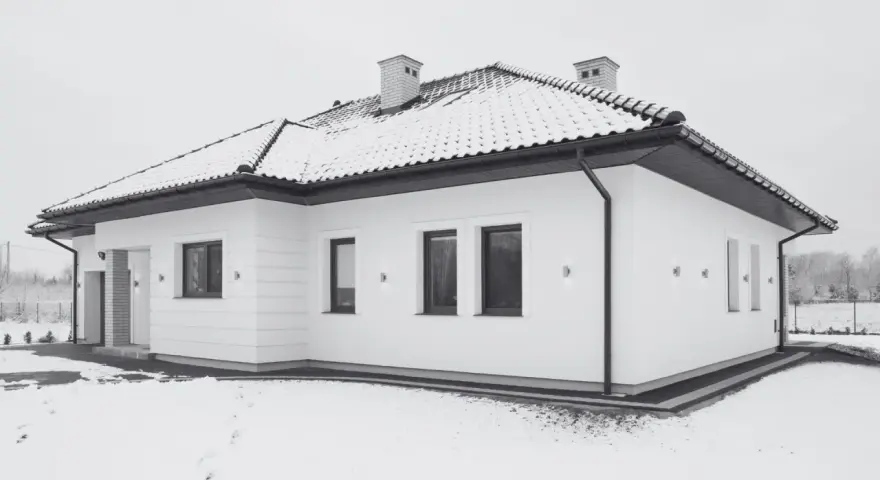 Nowoczesny, biały budynek wolnostojący z dachem pokrytym śniegiem. Wokół domu leży śnieg, tworząc zimową scenerię.