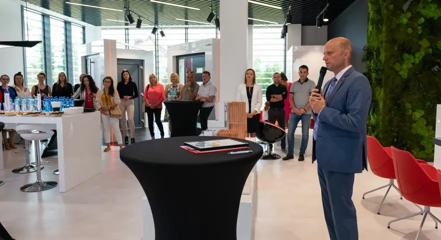 Mężczyzna w garniturze przemawia do grupy osób w nowoczesnym salonie okien i drzwi.