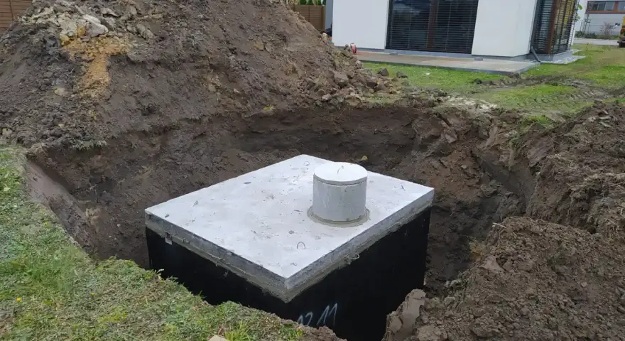 Gotowy odpływ z rynny do ziemi, betonowy zbiornik w wykopie, obok hałda ziemi i nowoczesny dom.