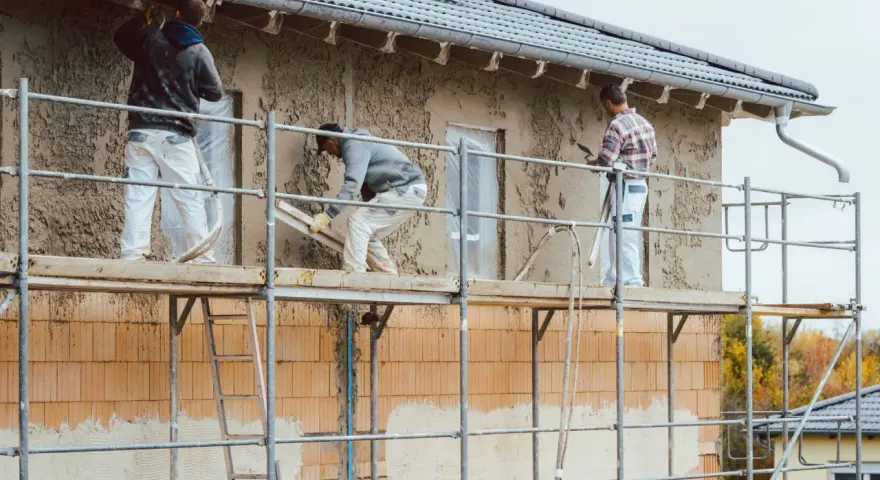 Pracownicy na rusztowaniu wykonują tynkowanie elewacji domu, nakładając zaprawę na ściany.