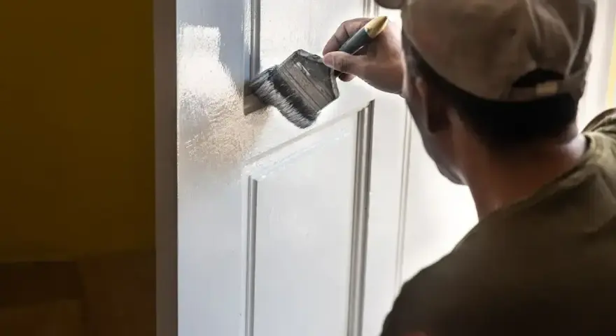 Mężczyzna w czapce z daszkiem maluje białe drzwi pędzlem. Odświeża wnętrze domu.