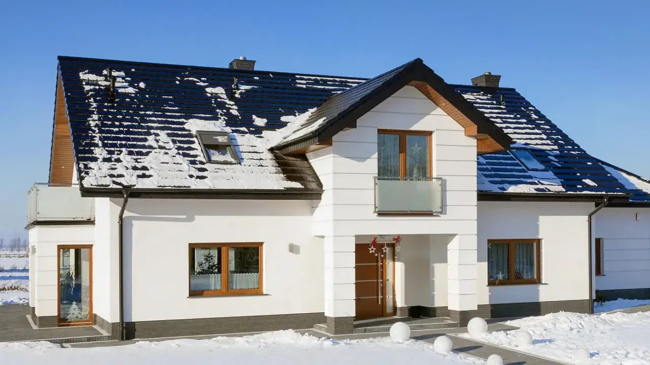 Nowy dom z białymi ścianami i czarnym, częściowo ośnieżonym dachem ceglastym. Wokół śnieg i zimowa aura.