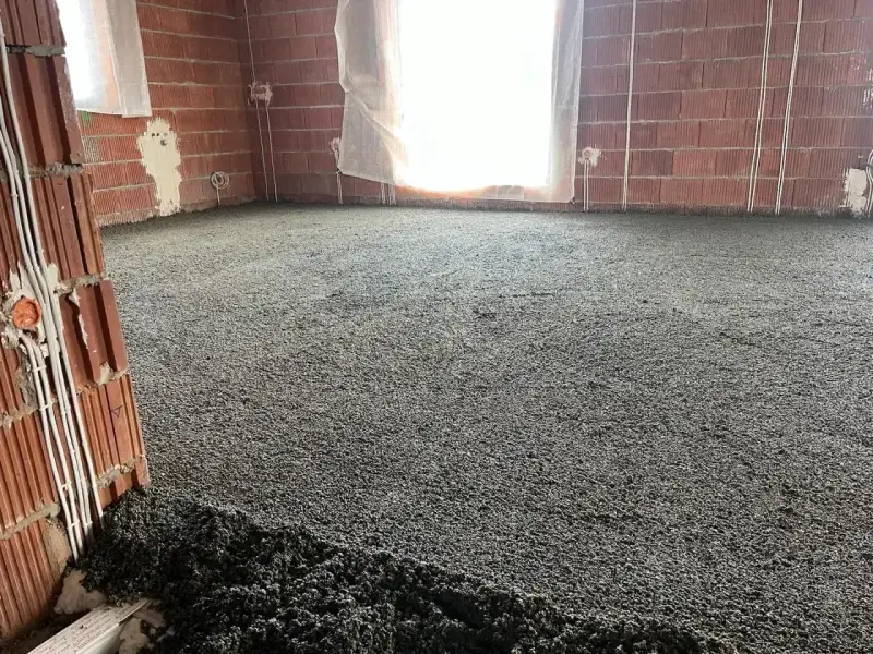 Wylewki styrobeton na podłodze w budowie. Ściany z cegły, okna zasłonięte, widoczne instalacje elektryczne.