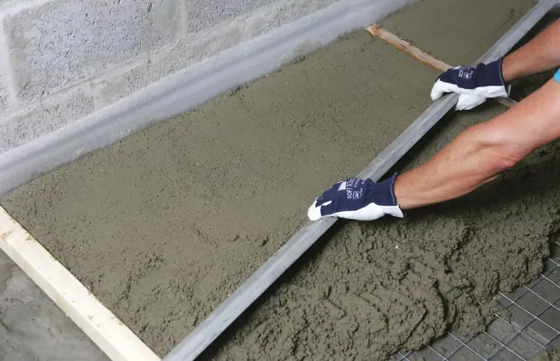 Równanie wylewki na ogrzewanie podłogowe. Ręce w rękawiczkach wyrównują świeży beton za pomocą łaty.